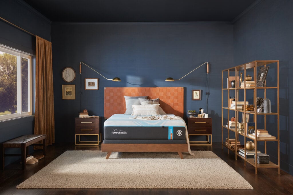 Tempur-Pedic LuxeBreeze Firm mattress displayed in a stylish bedroom setting with a textured headboard, decorative shelving, and warm lighting, highlighting its premium design and comfort features.