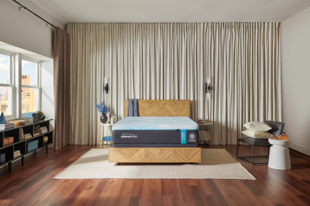 Tempur-Pedic LuxeBreeze Medium Hybrid mattress displayed in a stylish bedroom setting with wooden flooring, large windows, and elegant curtains, highlighting its cooling technology and pressure relief features.
