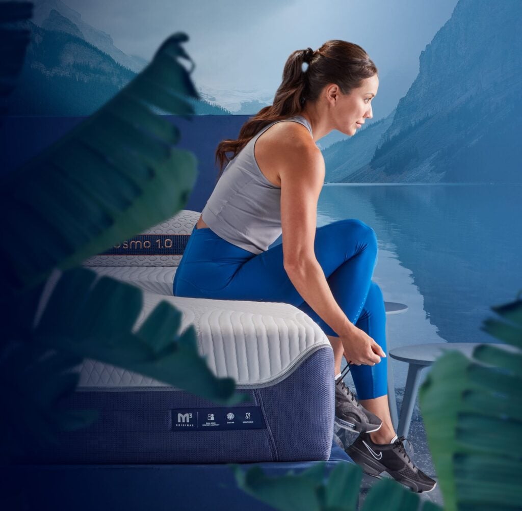 Woman sitting on Bedgear M3 modular mattress in activewear, preparing for exercise, with serene lake and mountains in the background, emphasizing personalized comfort and sustainable design.