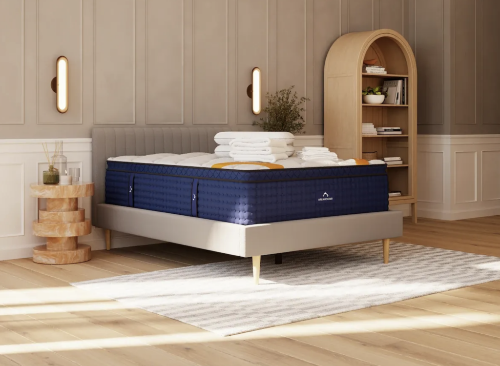 DreamCloud luxury hybrid mattress on a modern bed frame, surrounded by neatly stacked white towels, wooden nightstand with decorative items, and a stylish bookshelf in a cozy bedroom setting.