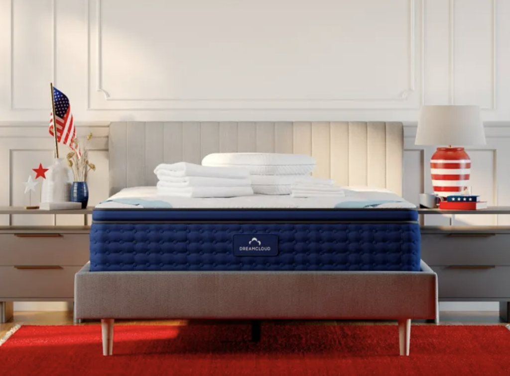 Luxury mattress setup with DreamCloud branding, featuring a plush blue mattress, neatly arranged white bedding, and decorative elements like an American flag and red-striped lamp in a stylish bedroom setting.