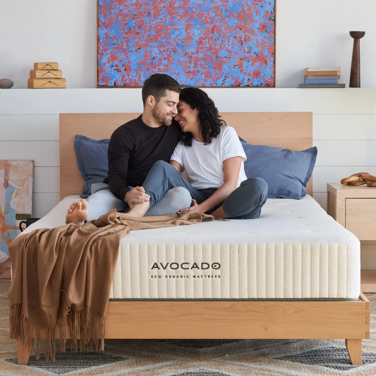 Couple sitting on Avocado Eco Organic mattress in a cozy bedroom, emphasizing comfort and quality sleep.