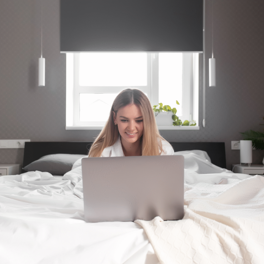 Young woman sitting on bed with laptop, smiling, in a modern bedroom, representing personalized mattress quiz experience at Midwest Mattress Company for better sleep.