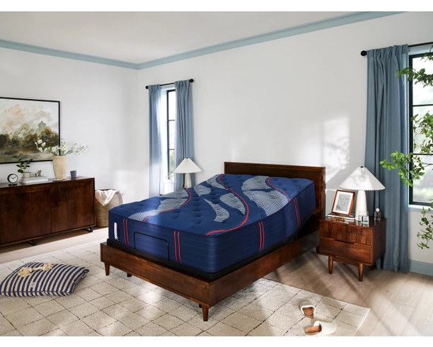 Sealy Brenham Ultra Firm TT mattress on wooden bed frame in a modern bedroom setting, featuring decorative pillows, bedside lamp, and natural light from windows.