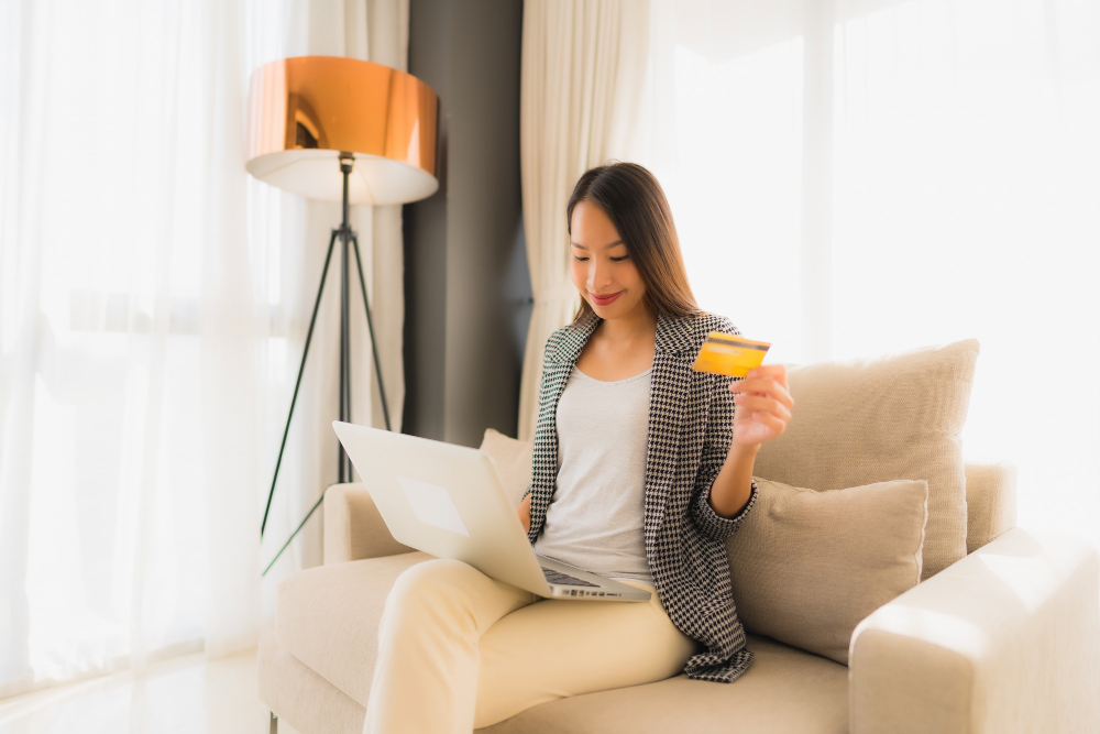 Woman sitting on a couch using a laptop and holding a credit card, symbolizing online shopping and flexible financing options for mattresses at Midwest Mattress Company.